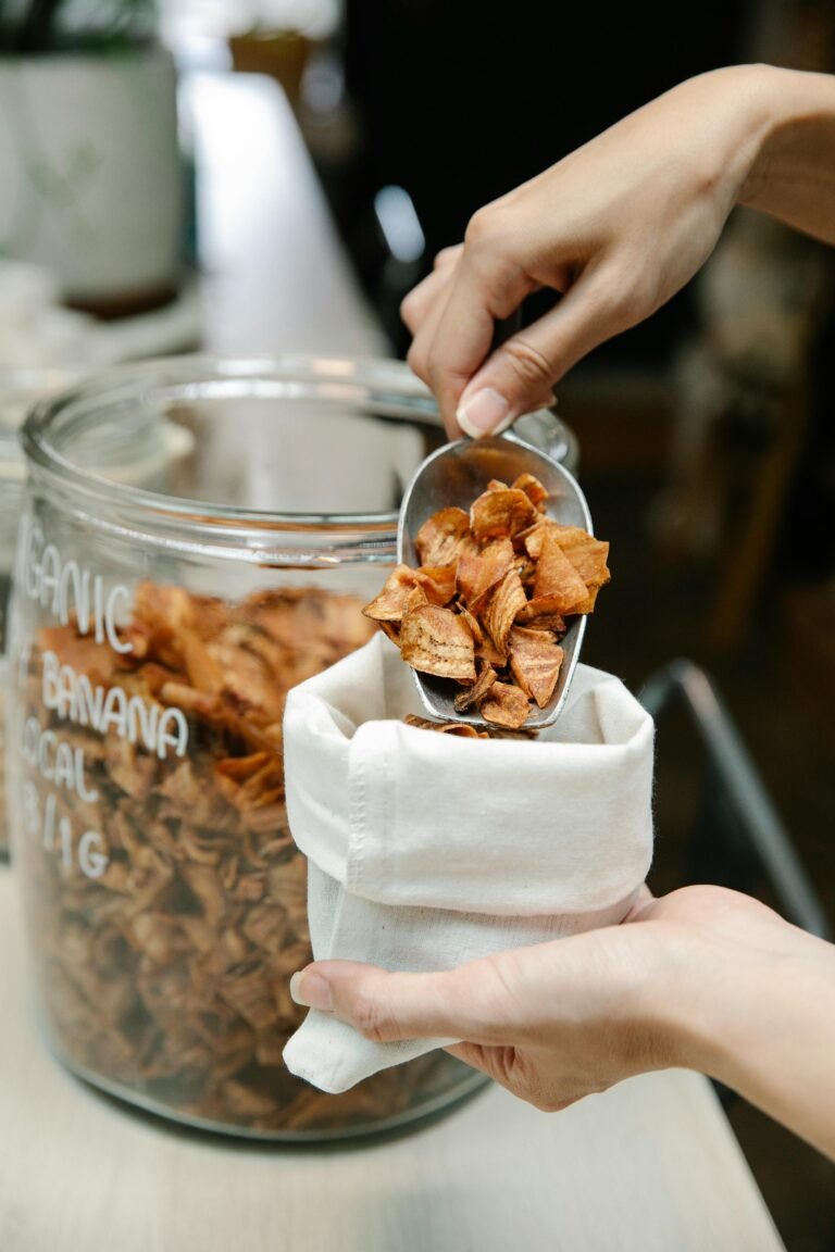 Close-up of organic banana chips being scooped from a glass jar into a cloth bag.
